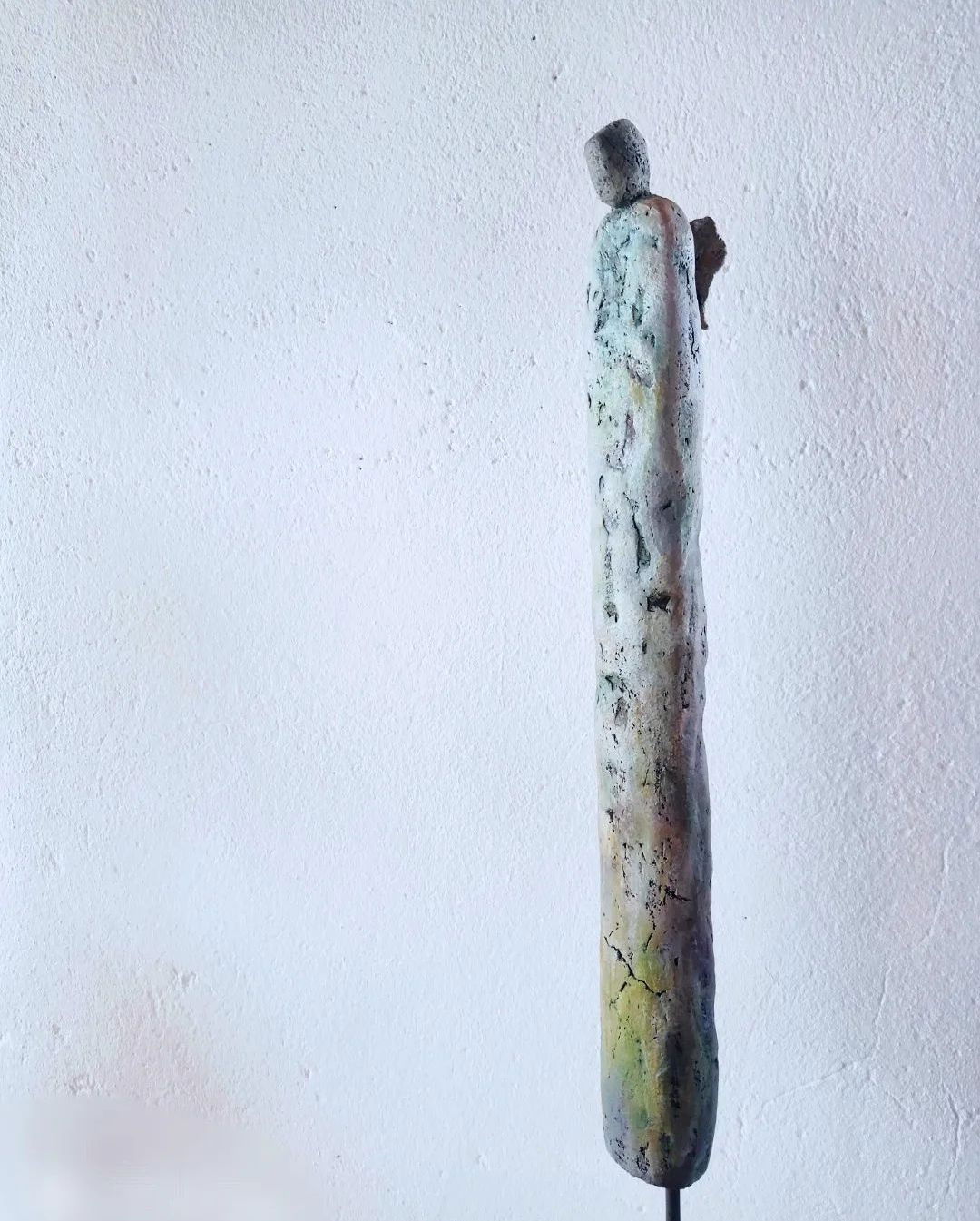 A tall, elongated ceramic figure stands in fragile solitude, its weathered patina and crackled surface embodying minimalism and serene abstraction against a textured backdrop. Artwork style is Figurativ and technique Keramik.