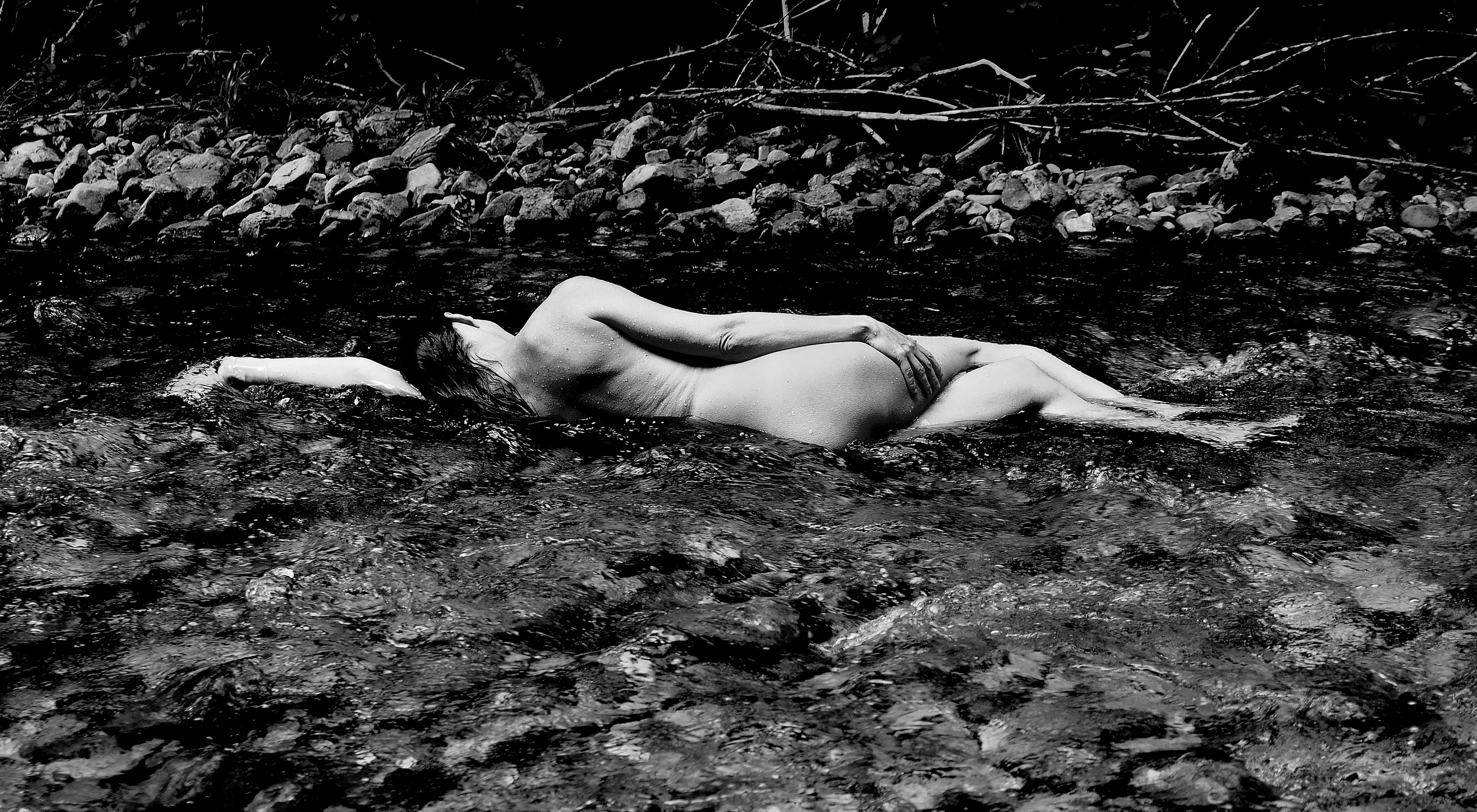 A monochromatic figurative artwork of a nude figure lying languidly in a peaceful stream, emphasizing tranquility, natural contrast, and ethereal solitude. Artwork style is Figurativ and technique Sublimationsdruck auf Leinwand.