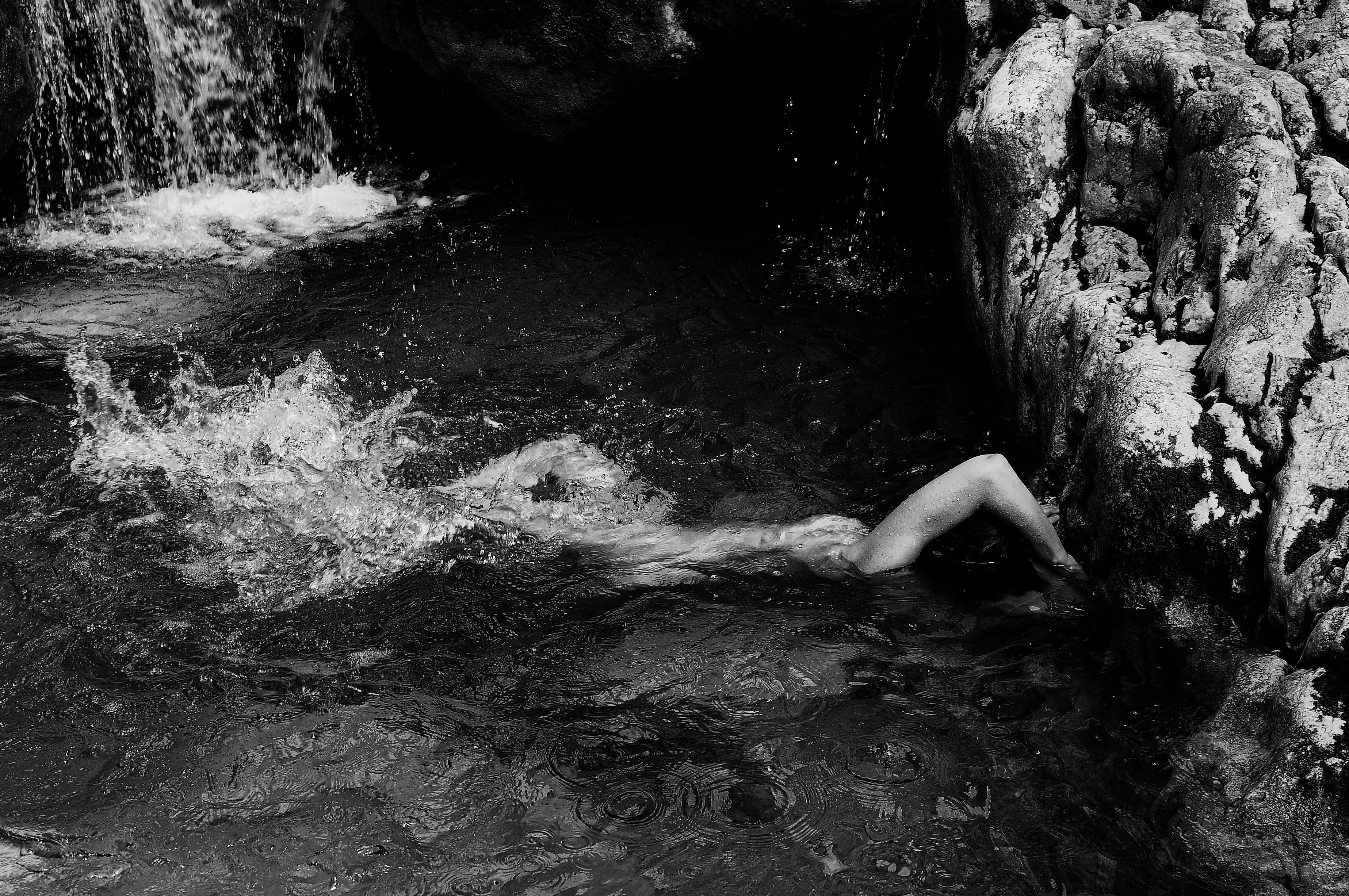 An evocative black-and-white artwork portraying a submerged figure in water by a rocky ledge, capturing the fluidity and stillness of nature's tranquil embrace. Artwork style is Figurativ and technique Sublimationsdruck auf Leinwand.