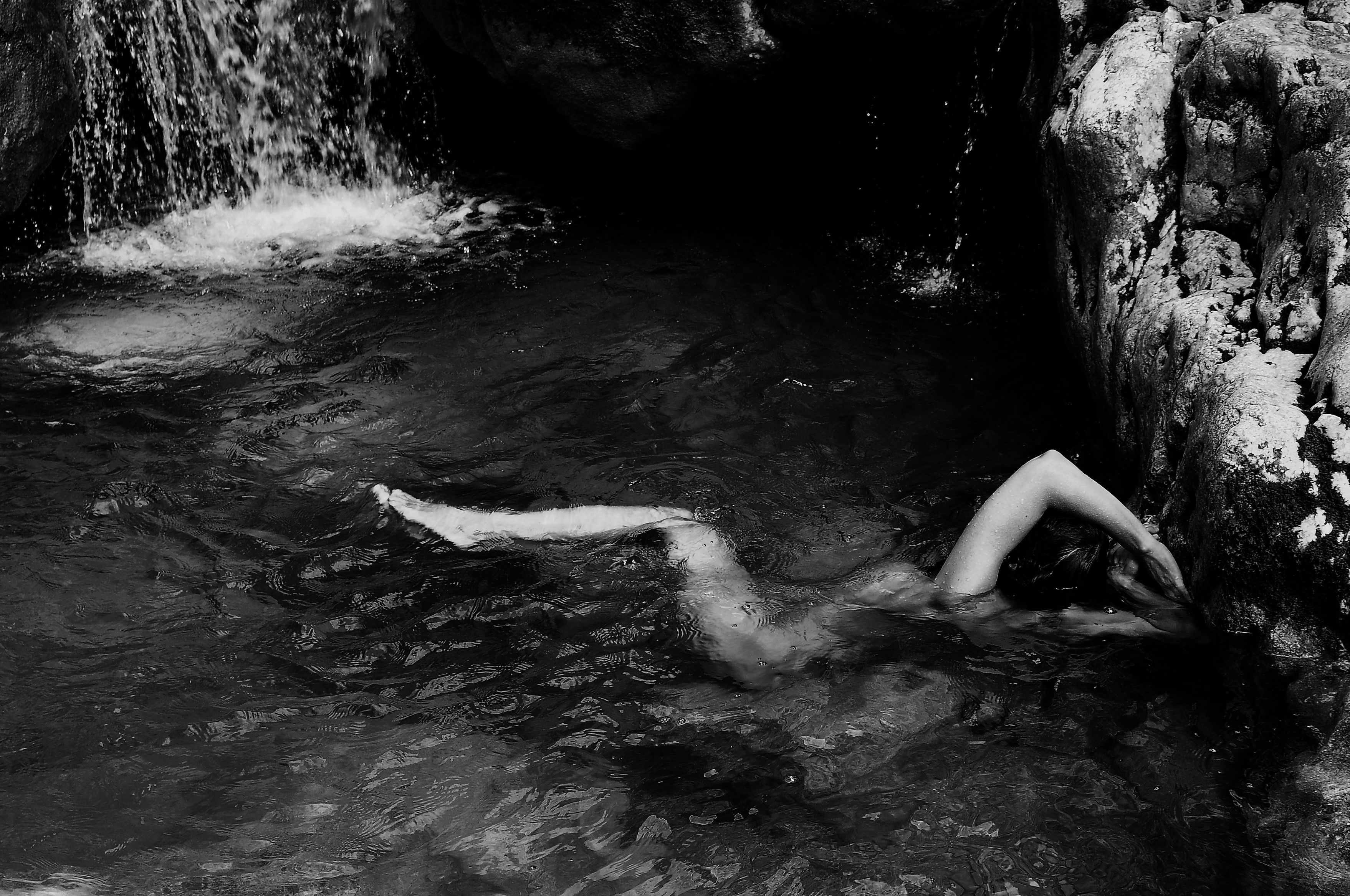 Grayscale artwork of a nude figure immersed in a serene waterfall pool, embracing the rocky surroundings, capturing the fluidity and tranquility of nature. Artwork style is Figurativ and technique Sublimationsdruck auf Leinwand.